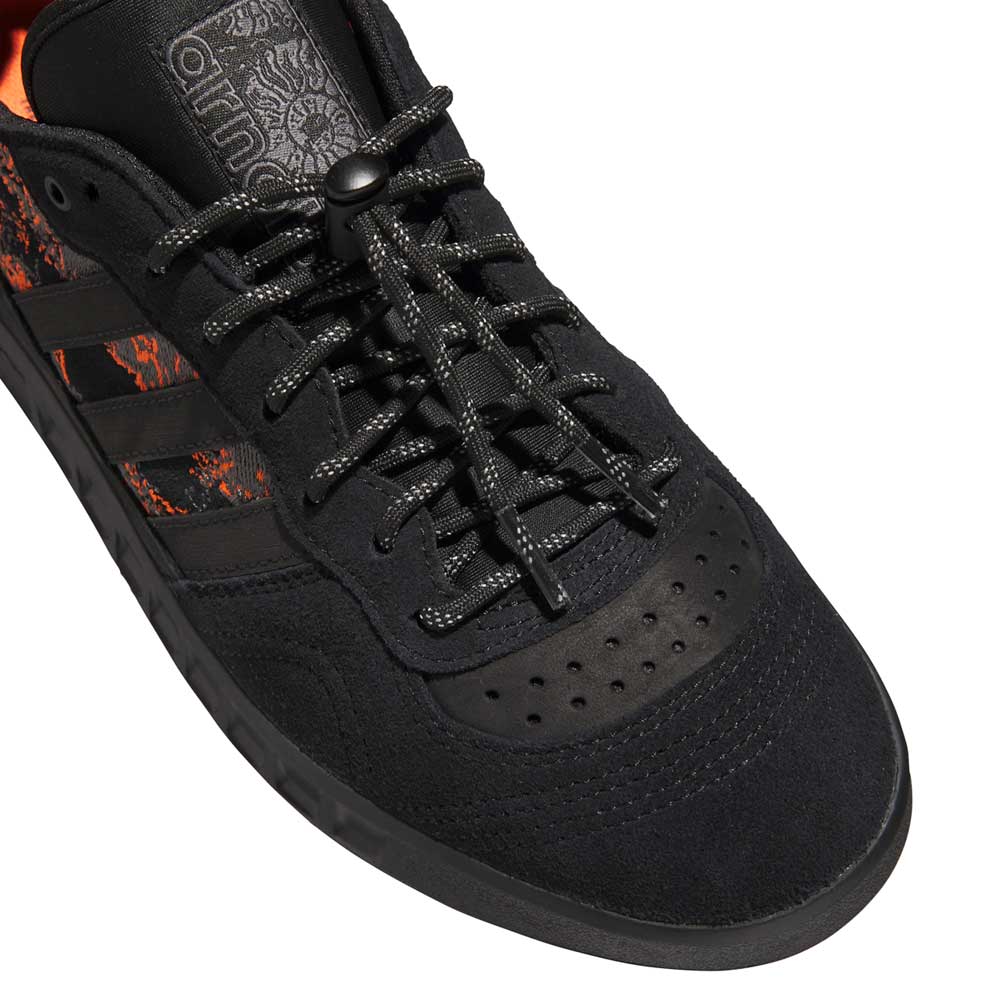 Close-up of the Handball Top x Mike Arnold Shoes in core black suede from adidas Skateboarding, featuring patterned laces, suede and leather textures, a subtle orange side accent, and a textured tongue with visible Adidas logo.