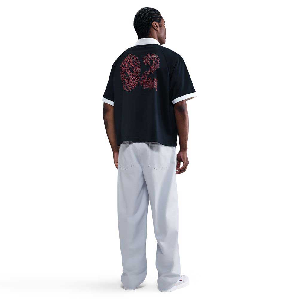 A person stands facing away, wearing a black short-sleeve shirt with “02” in red on the back, IB2992085 Skate Trousers Pant Loose Fit Straight Leg Nike SB Light Blue, and white sneakers against a plain white background.