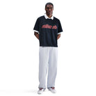 A person stands facing forward, wearing a black short-sleeve Nike SB shirt, IB2992085 Skate Trousers Pant Loose Fit Straight Leg Nike SB Light Blue, and white sneakers. Their hands are behind their back against a plain white background.