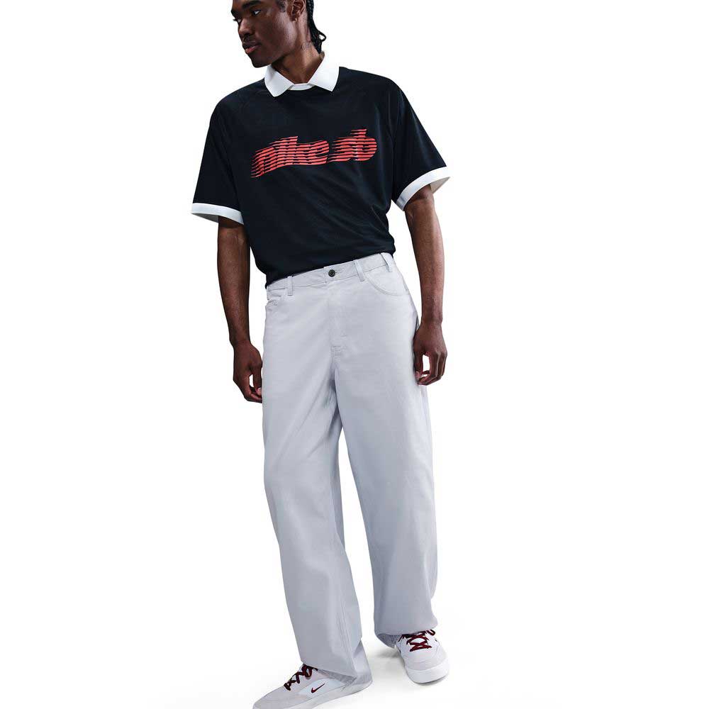 A man stands against a white background wearing a black Nike t-shirt with a red logo, white collared shirt underneath, loose fit straight leg Nike SB Light Blue skate trousers (IB2992085), and white sneakers with red accents.