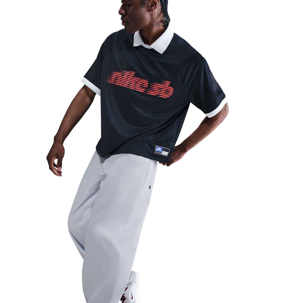 A person poses in a black short-sleeve shirt with a red, striped Nike logo and white trim, paired with IB2992085 Skate Trousers Pant Loose Fit Straight Leg Nike SB Light Blue and white sneakers against a plain white background.