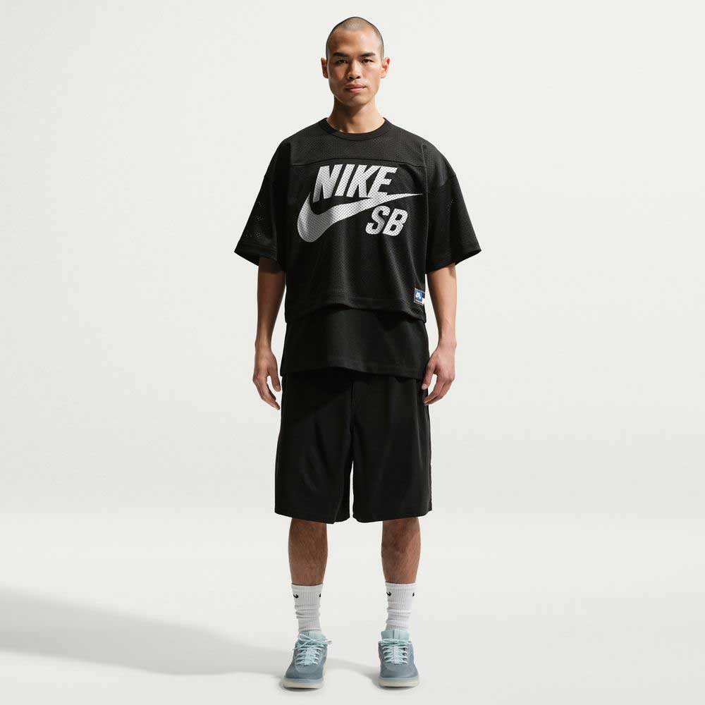 A person stands facing forward against a plain white background, wearing a loose black jersey, IF1096010 Black White Skate Shorts Nike SB, white socks, and light blue sneakers—perfect for a skate-ready fit.
