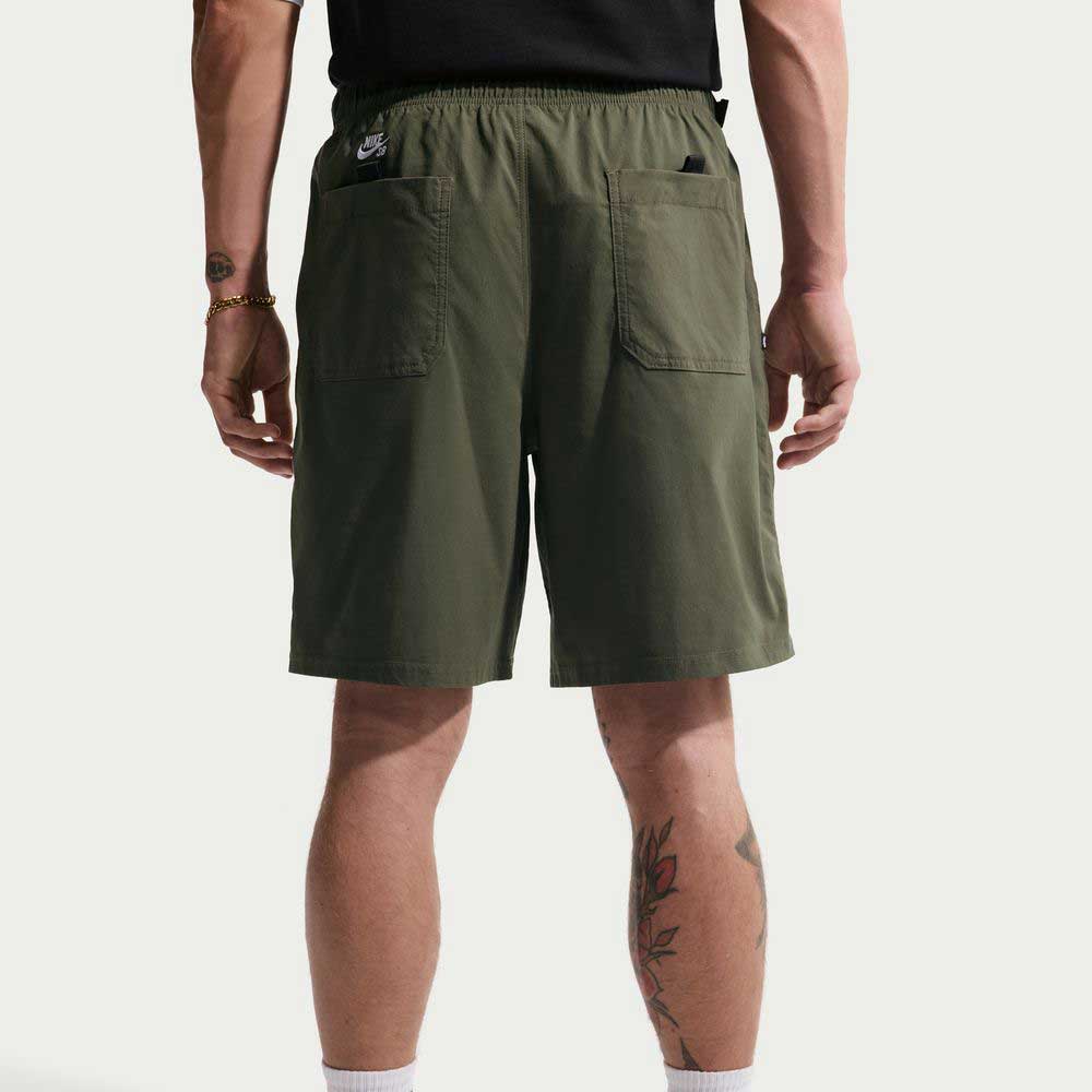 Wearing IF1096325 Cargo Khaki White Skate Shorts Nike SB with two back pockets and sweat-wicking fabric, a person in a black t-shirt and gold bracelet stands back to the camera, tattoos visible on left calf and right wrist.