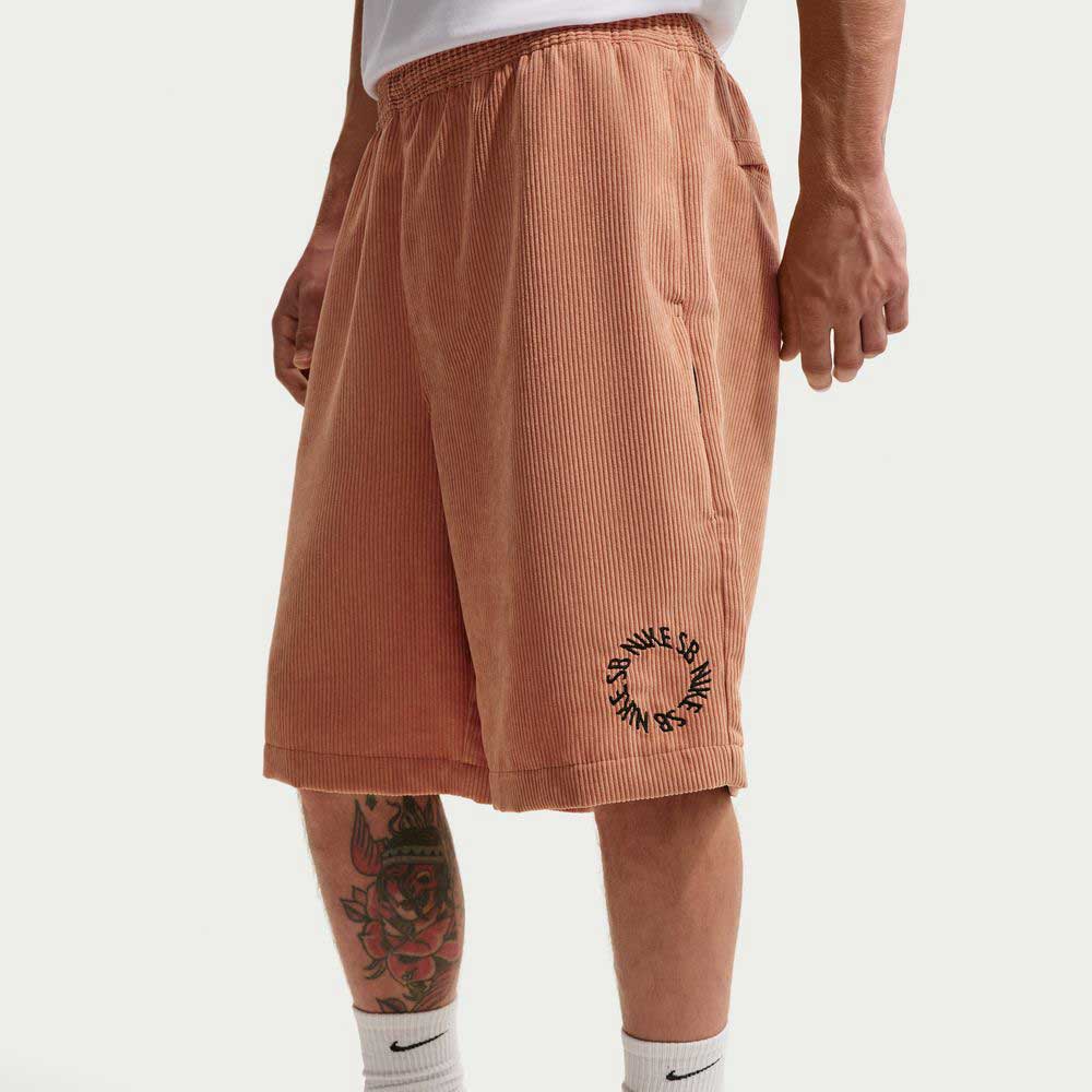 A person wearing Nike SB II4426217 Light Cognac Black corduroy skate shorts with “Save the Good Vibes” text, paired with a white shirt, white Nike socks, and visible leg tattoos.