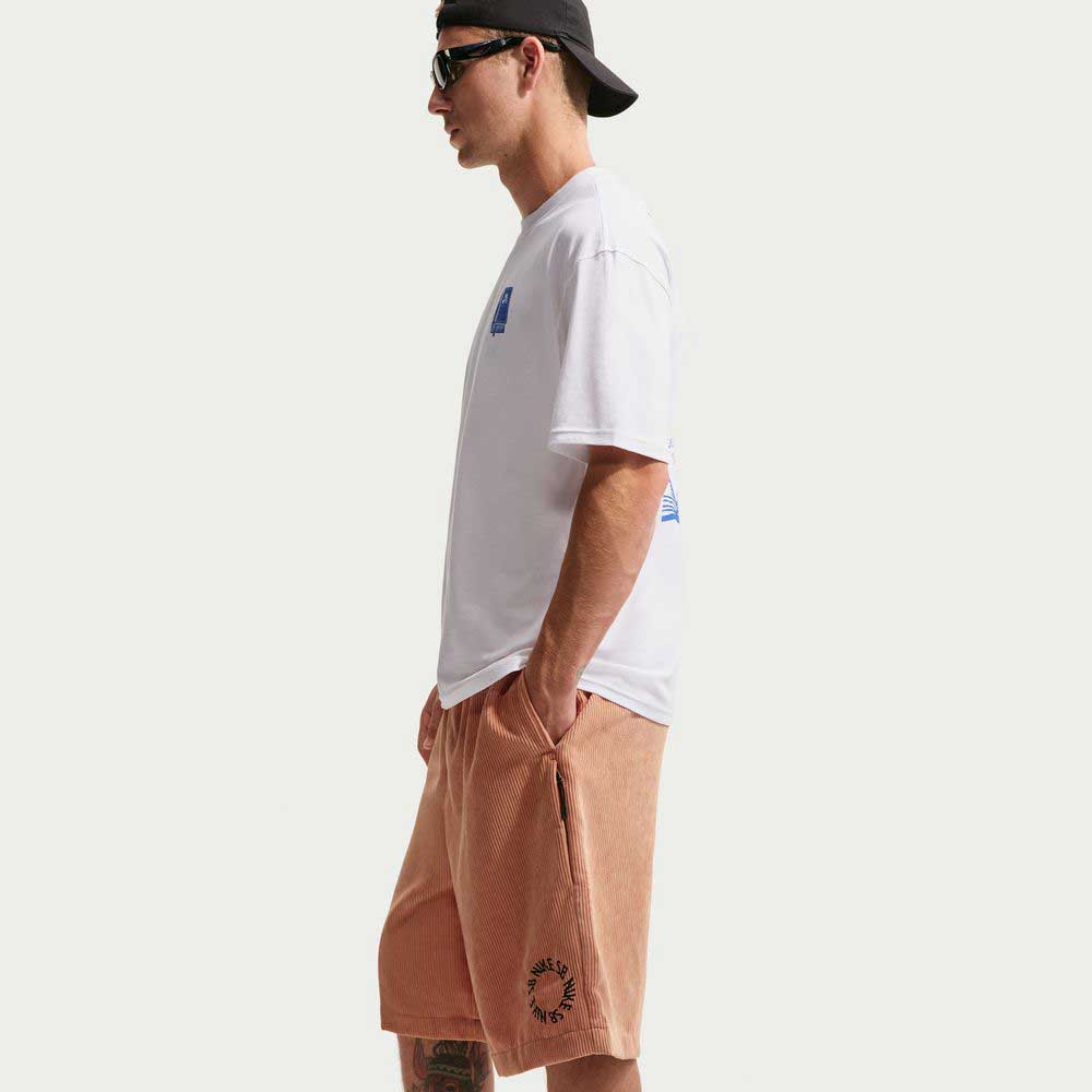 A man in sunglasses, a black backwards cap, a white t-shirt, and Nike SB II4426217 Light Cognac Black Corduroy Skate Shorts stands in profile against a plain white background.