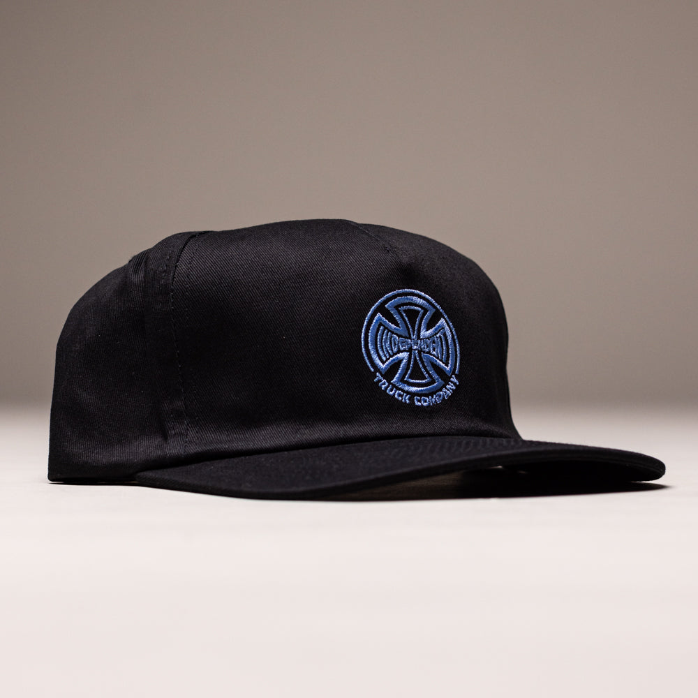 A black unstructured snapback hat with a curved brim and blue and white embroidered Independent Truck Co logo on the front, shown on a light surface. Product: Truck Co Legacy Black Hat Tan Snapback OS Unisex by Independent Skateboards.