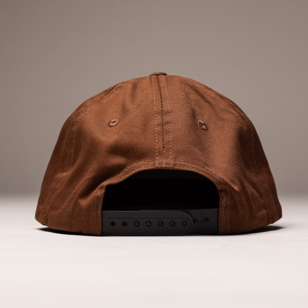 The Truck Co Legacy Brown Hat by Independent Skateboards is a tan, unstructured snapback with a black adjustable strap and low-profile fit, shown from the back on a neutral background. Unisex, OS.