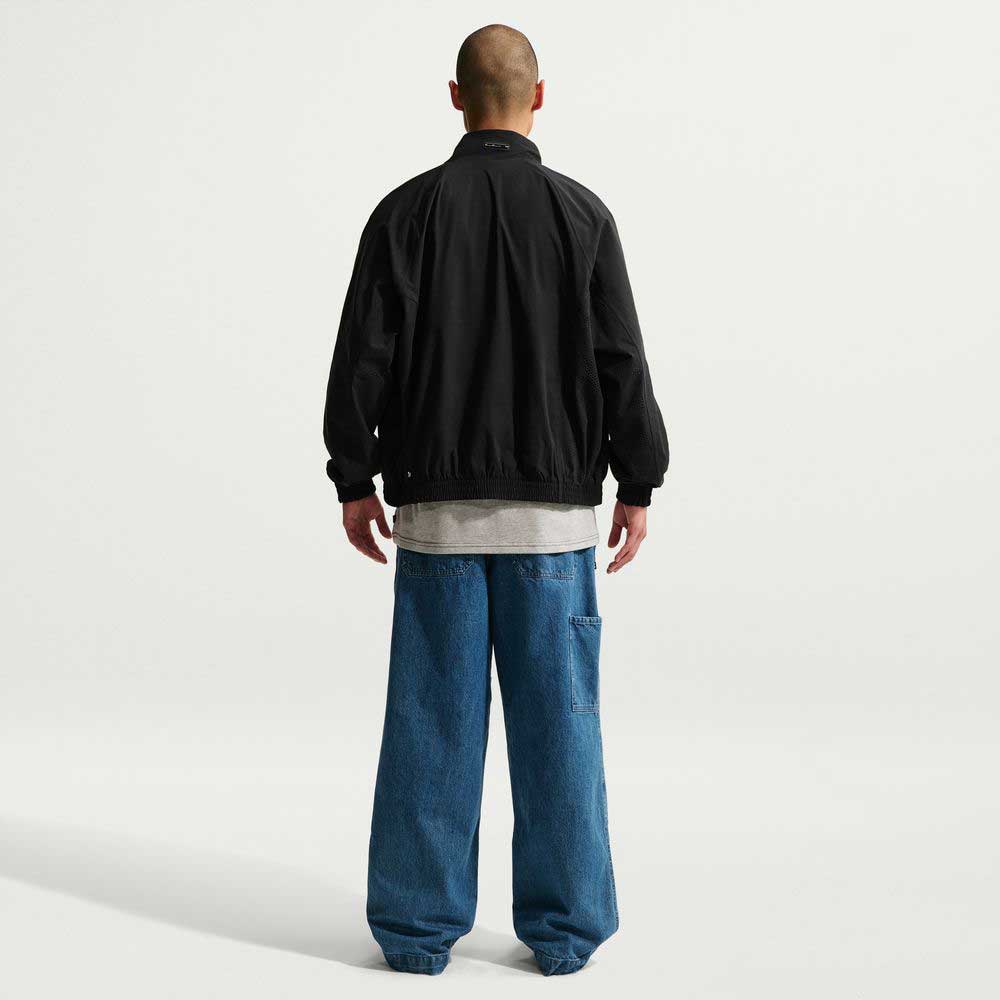 A person stands with their back to the camera, wearing a black Ishod Cargo Track Jacket Black IF0438010 Nike SB, a gray shirt, and loose blue jeans against a plain white background.
