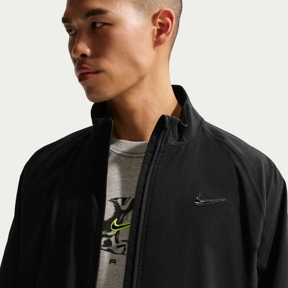 A person wears a black Nike SB Ishod Cargo Track Jacket (IF0438010) with a logo on the chest over a grey Nike t-shirt, looking to the side against a light background.