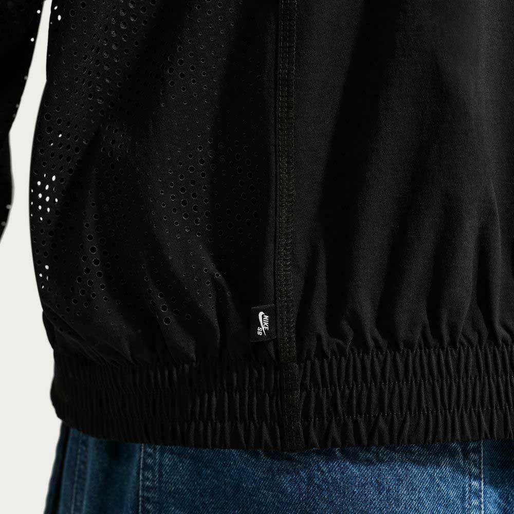 Close-up of a person wearing the Nike SB Ishod Cargo Track Jacket Black IF0438010 with perforated details, elastic waistband, and a small Nike tag, styled with blue denim jeans.