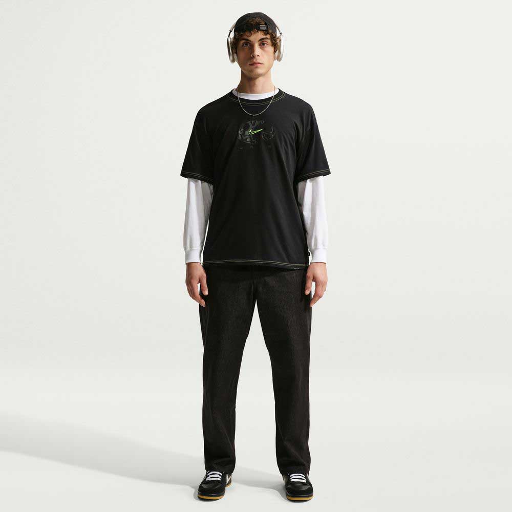 A person stands facing forward against a plain white background, wearing black pants, black sneakers, headphones, a long-sleeve white shirt, and the Ishod Tee Black IH1437010 Nike SB T-Shirt.