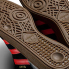 Close-up of the brown rubber soles of JH8134 Black Orange Gum Busenitz Adidas Skateboarding Suede Shoes, showcasing intricate tread patterns, the Adidas logo, and part of the shoe’s black suede side with orange stripes.