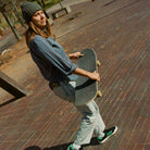 A person in a vintage beanie, plaid shirt, jeans, and JH8143 Nora Black Green Suede Adidas Skateboarding Shoes stands outdoors on a brick path, holding a skateboard and looking to the side.