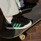 Wearing JH8143 Nora Black Green Suede Adidas Skateboarding Shoes, a person in light blue jeans stands on a black skateboard with white wheels on brick pavement.