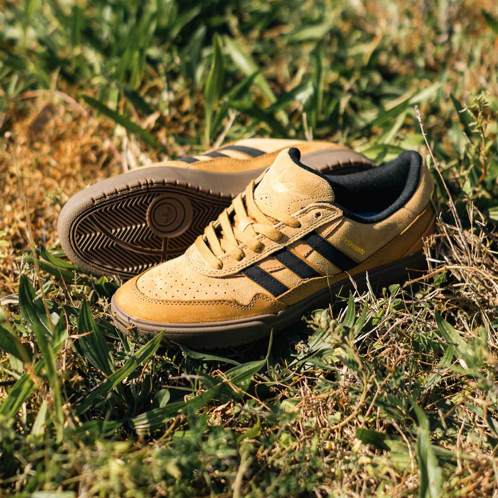 A pair of JI0851 Mesa Black Gum Tyshawn 2 II Adidas Leather Suede Skateboarding Shoes, tan with black stripes, rest on grass in sunlight, highlighting their cupsole construction from both side and top views.
