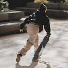 A person in a black hoodie and JM5832 Gray Clay Kader Sylla Pant Adidas is skateboarding outdoors on a sunny day, pushing off the ground with one foot while holding the skateboard with the other.