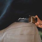 Close-up of a hand tucking in a dark shirt, featuring the JM5832 Gray Clay Kader Sylla Pant from Adidas, styled with a brown belt and a black leather "Kader" label on the waistband.