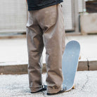 A person stands on a sidewalk holding a skateboard upright, showing the back of their legs in JM5832 Gray Clay Kader Sylla Pant Adidas with a straight-leg fit and black shoes.