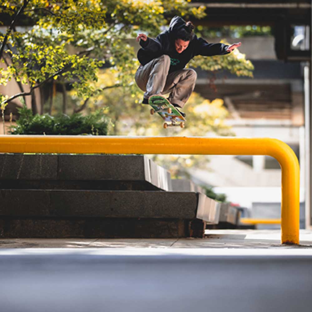 A skateboarder performs a trick over a yellow rail in the city, wearing JM5832 Gray Clay Kader Sylla Pant Adidas with a straight-leg fit amid trees and concrete structures.