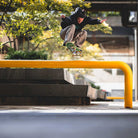 A skateboarder performs a trick over a yellow rail in the city, wearing JM5832 Gray Clay Kader Sylla Pant Adidas with a straight-leg fit amid trees and concrete structures.