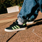 Close-up of a person in rolled-up blue jeans wearing JP5655 Black Neon Green Busenitz Vulc Suede Adidas Skateboarding Shoes, standing outdoors on a textured stone surface.