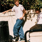 Two men in casual clothes and sneakers are outdoors by a stone wall. One leans while the other sits, both wearing JP5655 Black Neon Green Busenitz Vulc Suede Adidas Skateboarding Shoes. Sunlit tree shadows cross the scene; a trash can is on the left.