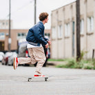 A young person in a blue jacket, beige cargo pants, and JP5861 Red White Gold Gazelle ADV Adidas Skateboarding Suede Shoes is skateboarding on an urban street with blurred buildings and cars in the background.