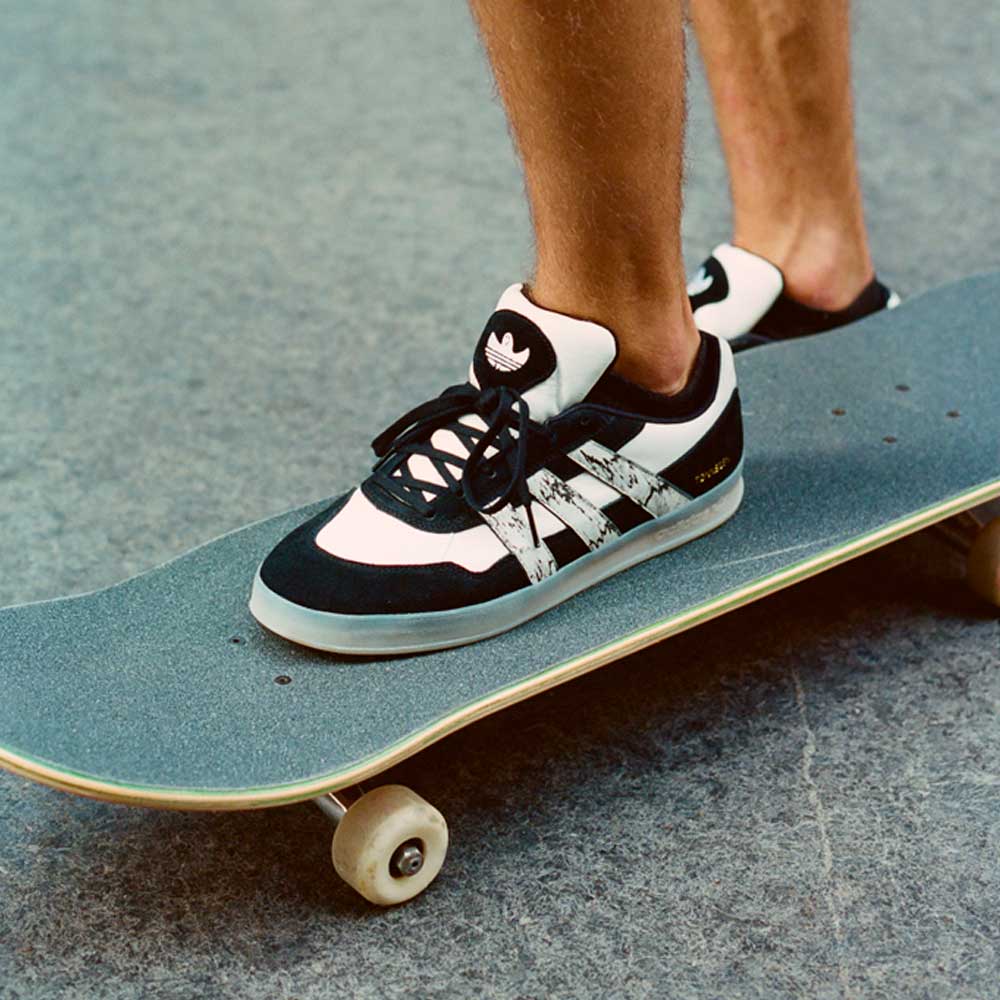 Close-up of a person wearing JR7101 Gustav Tonnesen Black White Aloha Super Suede Leather Skateboarding Shoes, standing on a skateboard, with only their lower legs and feet visible on a grey concrete surface.
