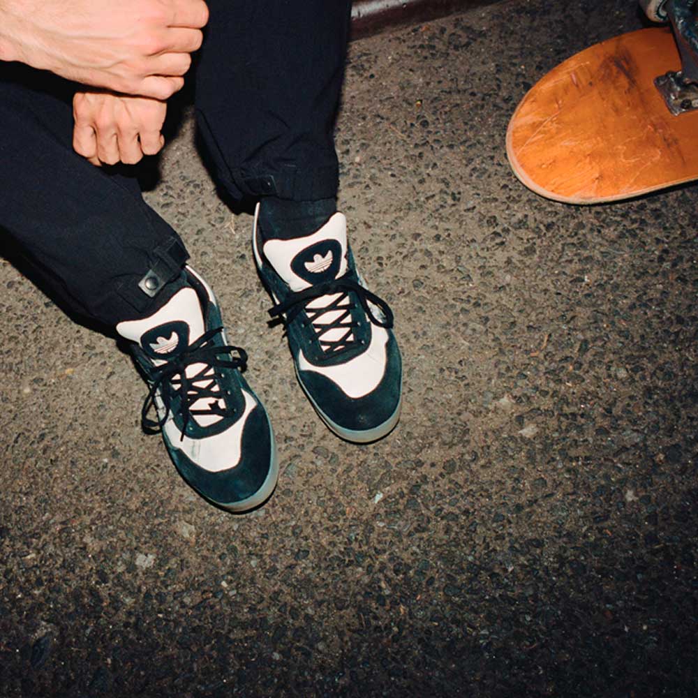 A person sits on asphalt with one hand on their knee, wearing JR7101 Gustav Tonnesen Black White Aloha Super Suede Leather Skateboarding Shoes. Part of a skateboard and their lower legs are visible, all lit with a flash.