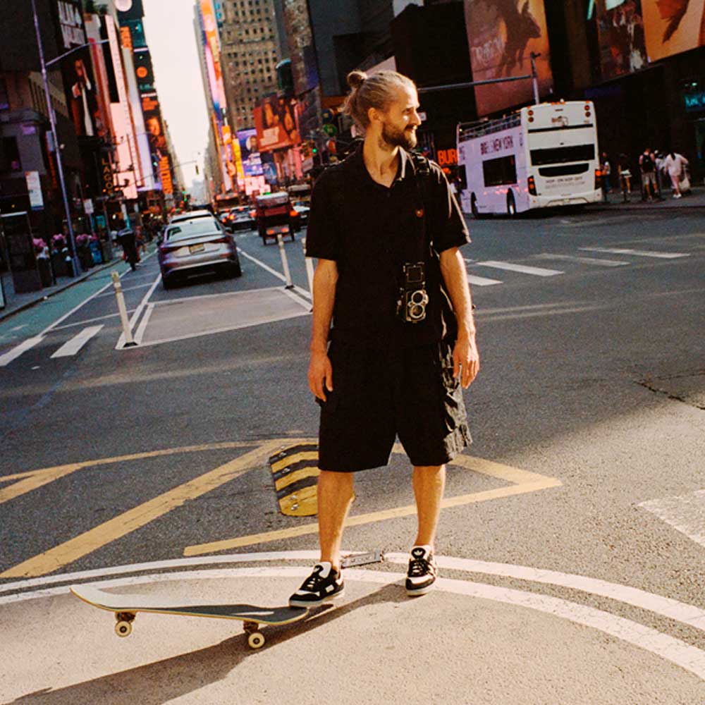 A bearded man with long hair in a bun stands on a skateboard in the city, smiling in black clothes with a camera. He sports JR7101 Gustav Tonnesen Black White Aloha Super Suede Leather Skateboarding Shoes, ready to capture every moment.