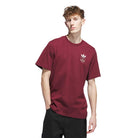A young man with light skin and short, curly brown hair wears a burgundy JW0056 Skate Tee from Adidas Skateboarding with a small white Adidas logo on the chest and black pants, standing against a white background. This tee is imported for comfort.