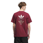 A person with short, light brown hair is seen from behind wearing the JW0056 Skate Tee Burgundy from Adidas Skateboarding, featuring three large beige and gray trefoil logos stacked on the back.