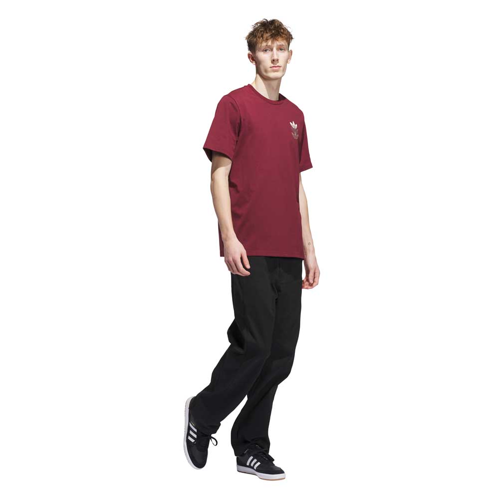 A person wearing a burgundy JW0056 Skate Tee from Adidas Skateboarding, black pants, and black Adidas sneakers stands against a white background. The imported tee has a small white logo on the chest.