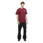 A young man stands against a white background, wearing a burgundy Adidas Skateboarding JW0056 Skate Tee with a small white logo on the chest, black pants, and black sneakers.