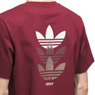 Person wearing the JW0056 Skate Tee in Burgundy by Adidas Skateboarding, featuring three large Trefoil logos in varying styles and shades on the back, with an Adidas Skateboarding logo near the hem. Made from imported fabric for lasting comfort.