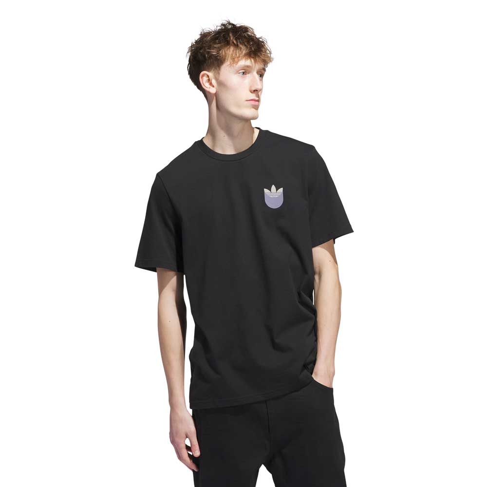 A young man with short, messy hair wears the JW0058 Skate Tee Black Olive Strata by Adidas Skateboarding, featuring a small multicolored logo on the left chest, paired with black pants. He stands against a white background, looking to the side.
