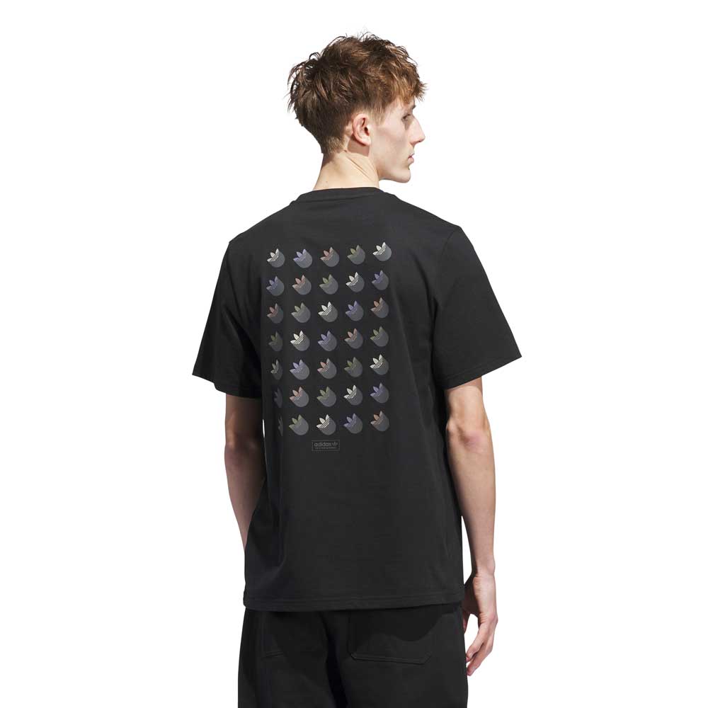 A young man with short, light brown hair wears the JW0058 Skate Tee Black Olive Strata from Adidas Skateboarding, featuring a shiny trefoil logo grid on the back. He stands facing away, glancing left.