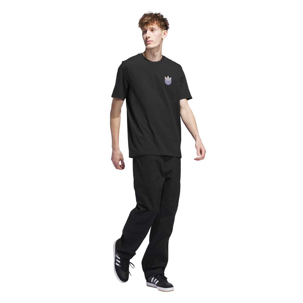 A young man stands against a white background, wearing a black JW0058 Skate Tee from the Black Olive Strata Adidas Skateboarding collection, paired with black pants and black adidas sneakers with white stripes.