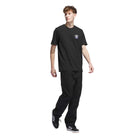 A young man stands against a white background, wearing a black JW0058 Skate Tee from the Black Olive Strata Adidas Skateboarding collection, paired with black pants and black adidas sneakers with white stripes.
