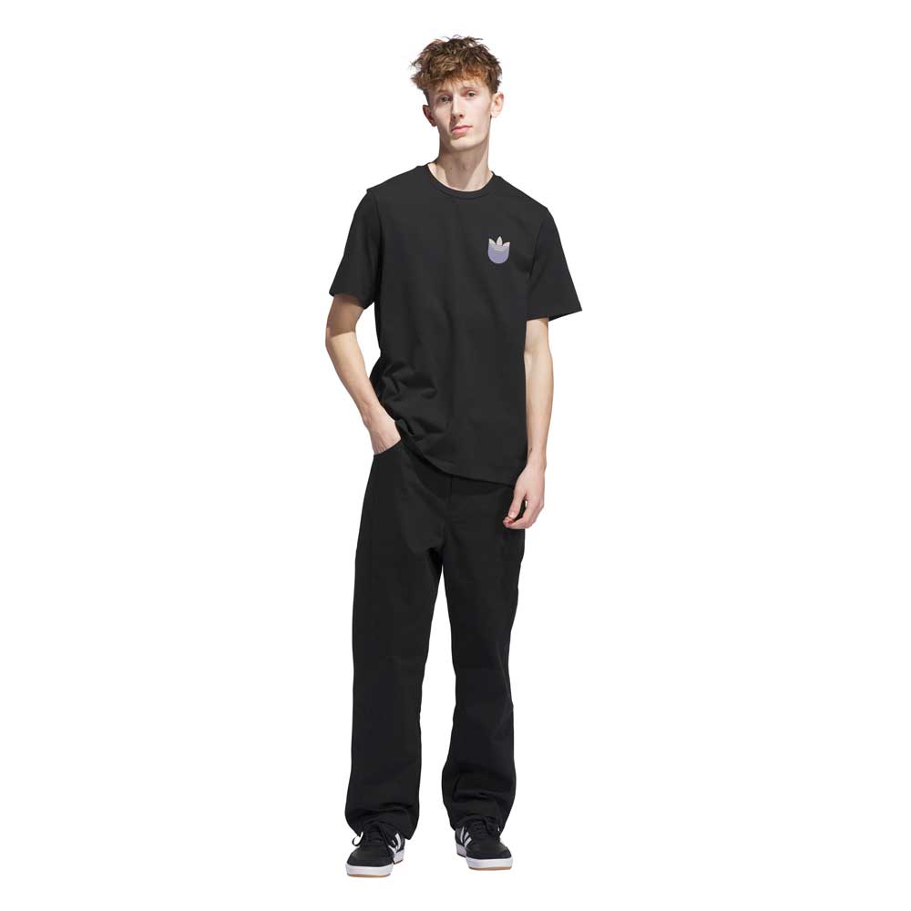 A young man stands against a white background wearing the JW0058 Skate Tee Black Olive Strata from Adidas Skateboarding, paired with black pants and black sneakers with white soles for a sleek, modern look.