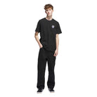 A young man stands against a white background wearing the JW0058 Skate Tee Black Olive Strata from Adidas Skateboarding, paired with black pants and black sneakers with white soles for a sleek, modern look.