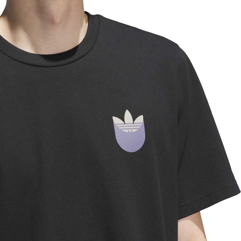 A person wears the JW0058 Skate Tee in Black Olive Strata by Adidas Skateboarding, featuring a light beige and lavender Trefoil logo on the left chest. Only their lower face, neck, and upper torso are shown. Imported.