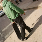 A person in a green hoodie and JX6827 Black Kader Sylla Pant Adidas walks on a street, holding a skateboard by their side. Their face isn't visible, and their shadow stretches across the pavement.