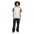 A person with curly hair wears a white T-shirt with brown and white striped sleeves, black JX6827 Black Kader Sylla Pant Adidas, and black sneakers, standing against a plain white background.