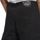 Close-up of a person wearing the JX6827 Black Kader Sylla Pant by Adidas, featuring cotton twill fabric, two back pockets, Trefoil logo on the right pocket, a black Adidas waistband label, and a tucked-in white shirt.