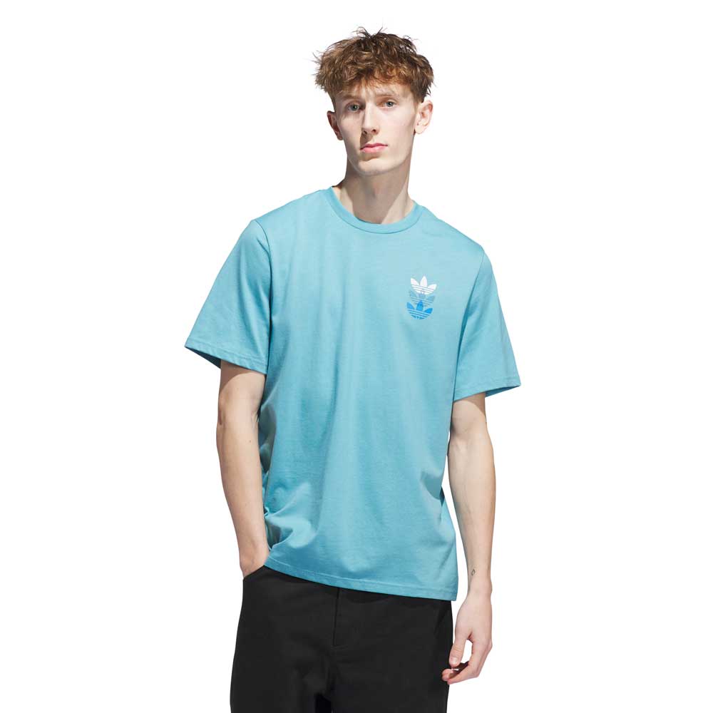 A young man wears a JY3954 Skate Tee in Preloved Blue Bright Royal from Adidas Skateboarding, featuring a small white chest logo, paired with black pants, against a plain white background.