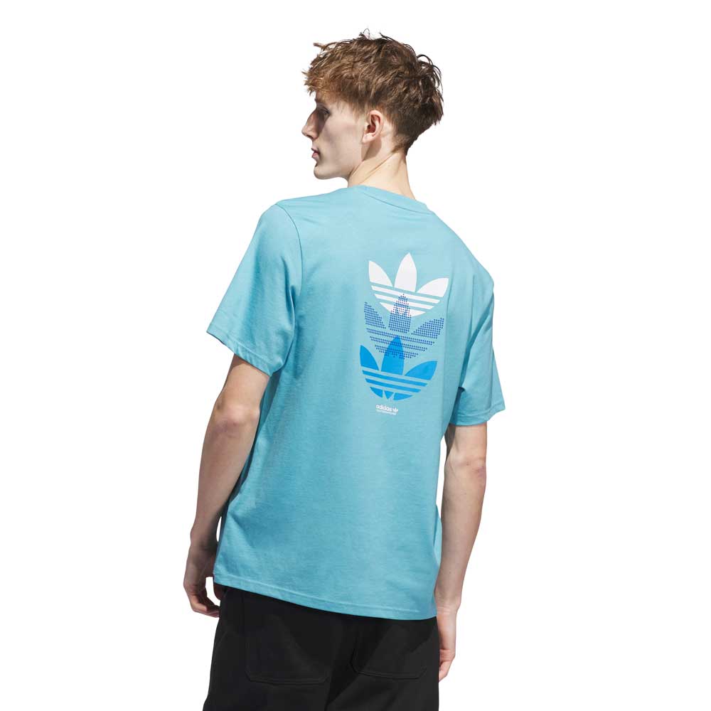 A young man with short brown hair faces away, modeling the Adidas Skateboarding JY3954 Skate Tee in Preloved Blue and Bright Royal, which features three trefoil logos on the back. He stands against a plain white background.
