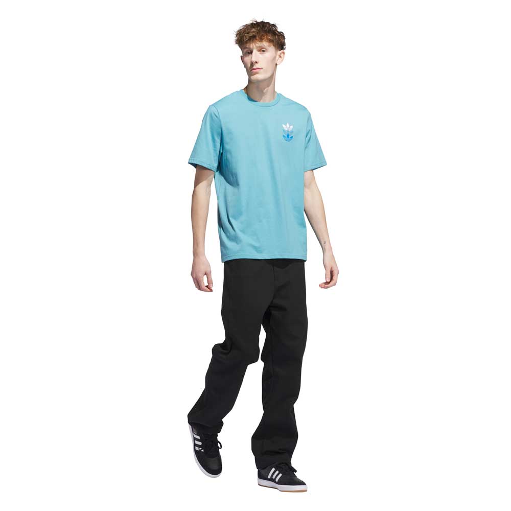 A young man with curly hair wears the JY3954 Skate Tee Preloved Blue Bright Royal from Adidas Skateboarding, black pants, and black Adidas sneakers with white stripes, standing against a plain white background.