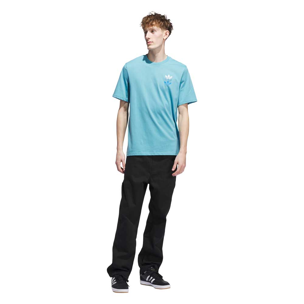 A young man stands against a white background wearing the JY3954 Skate Tee Preloved Blue Bright Royal Adidas Skateboarding shirt, black pants, and black Adidas sneakers. He looks slightly left with a neutral expression.