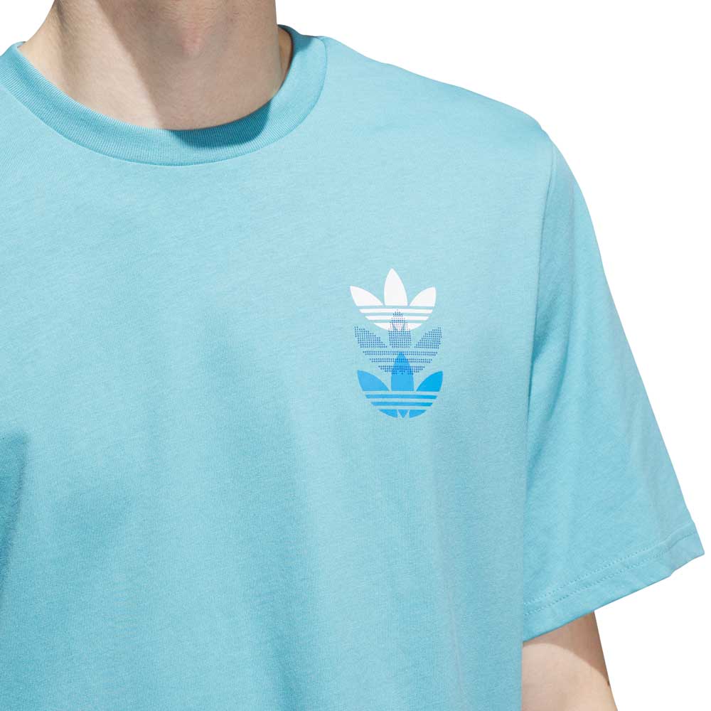 A person wearing the JY3954 Skate Tee in Preloved Blue/Bright Royal by Adidas Skateboarding, featuring three stacked trefoil logos in white and Preloved Blue on the left chest. The face is not visible.